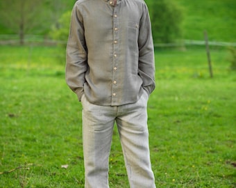 May include: A man wearing a light brown linen shirt with a mandarin collar and a button-up front. He is also wearing light gray linen pants and is standing in a grassy field.