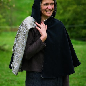 May include: A black hooded cape featuring a contrasting interior lining with a metallic silver and sage green floral damask pattern. The cape is worn over a brown long-sleeved top and a dark gray skirt, draping over the shoulders.