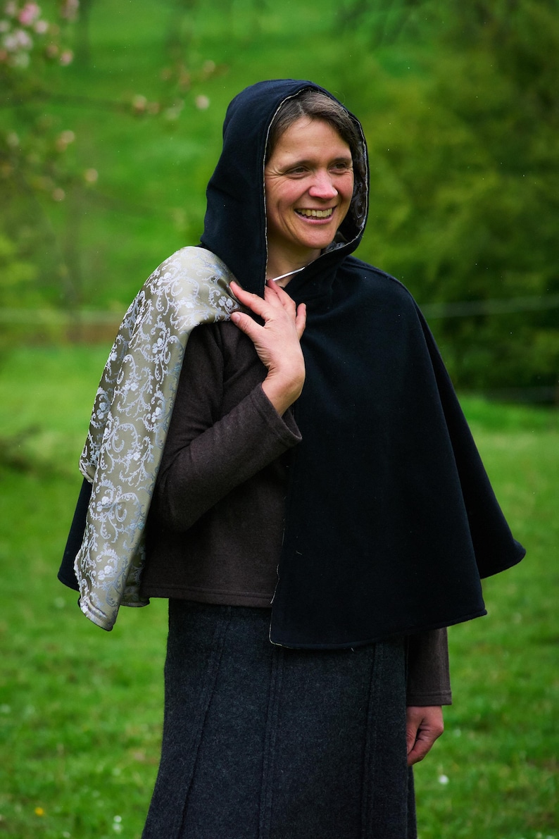 May include: A black hooded cape featuring a contrasting interior lining with a metallic silver and sage green floral damask pattern. The cape is worn over a brown long-sleeved top and a dark gray skirt, draping over the shoulders.