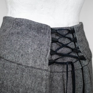 May include: Close-up of a gray herringbone skirt with a black lace-up detail at the back. The skirt has a high waist and a pleated design. The fabric appears to be a wool blend.