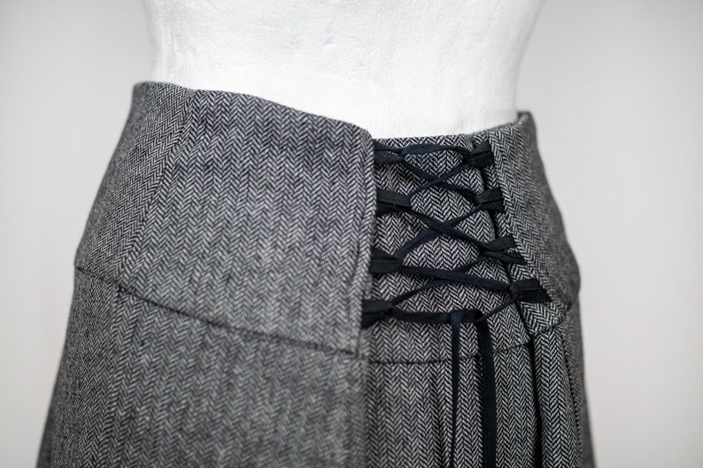 May include: Close-up of a gray herringbone skirt with a black lace-up detail at the back. The skirt has a high waist and a pleated design. The fabric appears to be a wool blend.