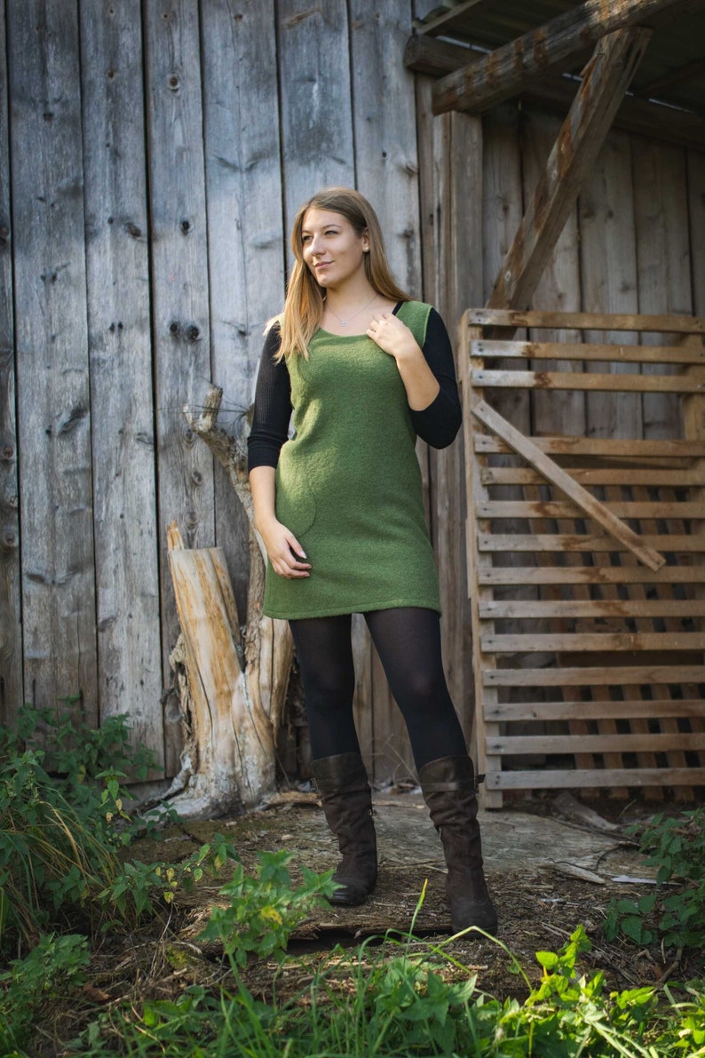 Puede incluir: Una mujer con un vestido de su&eacute;ter verde, medias negras y botas marrones. Est&aacute; de pie frente a una pared de madera y una puerta de madera.