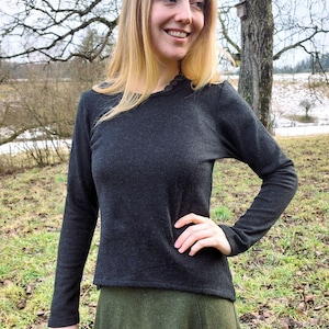 Puede incluir: Una camisa de manga larga gris oscuro con cuello redondo y una falda verde. La camisa tiene un delicado ribete de encaje alrededor del escote. La persona está de pie afuera frente a un árbol.