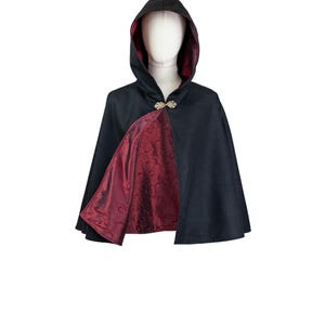 May include: A black hooded cape featuring a deep burgundy lining with a subtle tonal floral pattern. The garment is secured at the neck with an ornate gold-toned metal clasp, showcasing a structured hood and a flared silhouette.