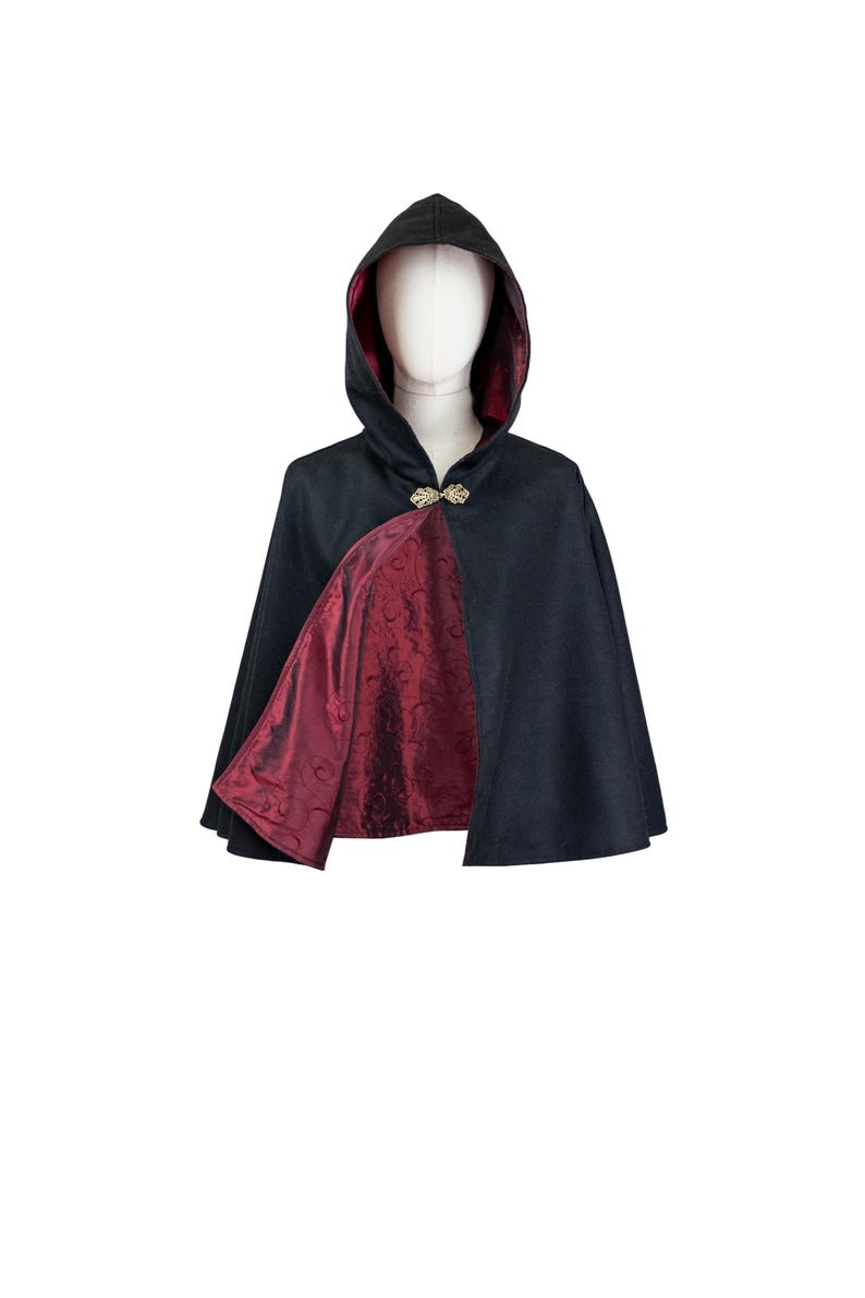 May include: A black hooded cape featuring a deep burgundy lining with a subtle tonal floral pattern. The garment is secured at the neck with an ornate gold-toned metal clasp, showcasing a structured hood and a flared silhouette.