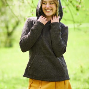 May include: A woman wearing a dark gray hooded sweater with a mustard yellow skirt. She is standing in a grassy field with trees in the background.