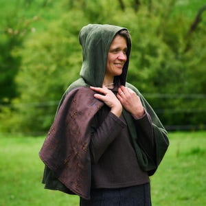 May include: A dark green hooded cloak featuring a brown interior lining with a subtle floral embroidery pattern. The garment is worn over a dark brown long-sleeved top and a dark gray skirt, providing full coverage for the upper body.