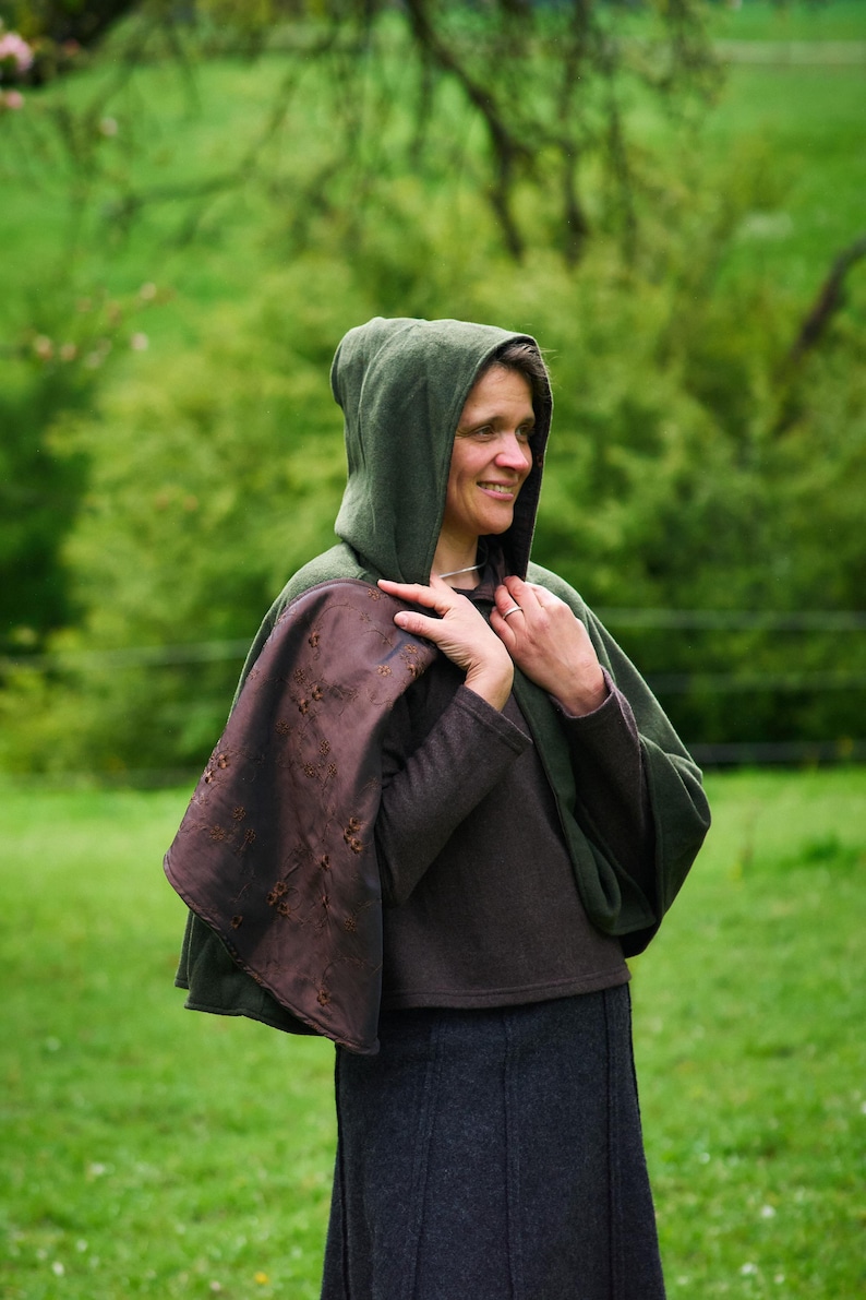 May include: A dark green hooded cloak featuring a brown interior lining with a subtle floral embroidery pattern. The garment is worn over a dark brown long-sleeved top and a dark gray skirt, providing full coverage for the upper body.