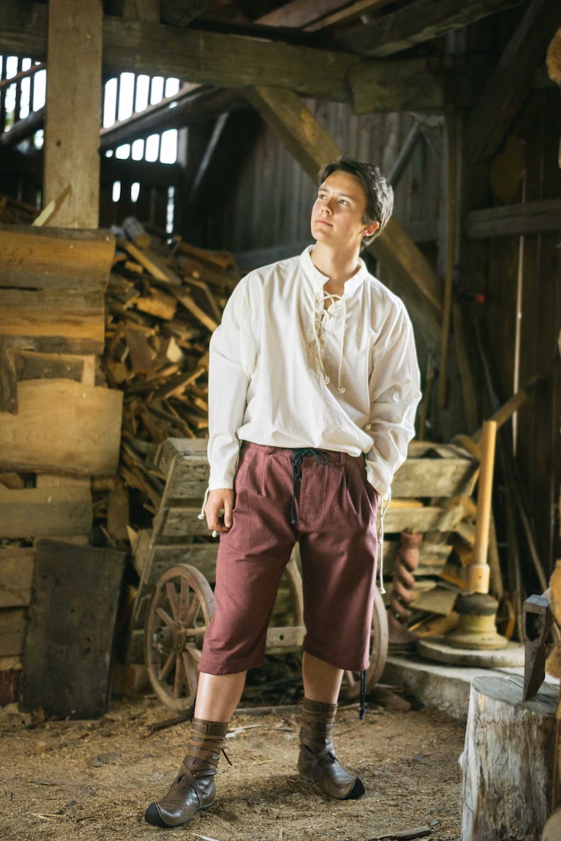 May include: A man wearing a white collared shirt with a lace-up neckline and burgundy shorts. He is standing in a rustic wooden barn setting.