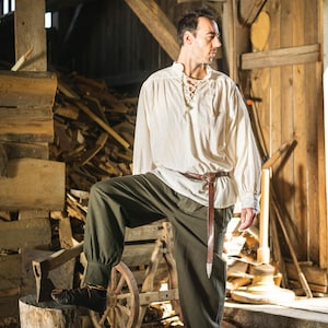 Op de afbeelding: Een man in een wit shirt met een kanten halslijn en een bruine broek staat in een houten schuur. Hij houdt een houten bijl in zijn rechterhand en kijkt naar rechts. De schuur is gevuld met houtresten en een houten kar.