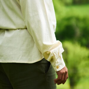 Op de afbeelding: Een close-up van een persoon die een wit linnen shirt draagt met een lange mouw die is opgerold en een gouden knoop. De persoon draagt een donkergroene broek.