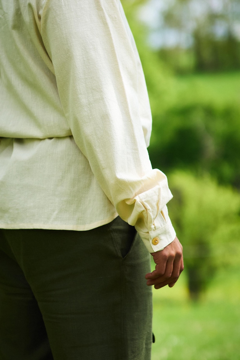 Op de afbeelding: Een close-up van een persoon die een wit linnen shirt draagt met een lange mouw die is opgerold en een gouden knoop. De persoon draagt een donkergroene broek.
