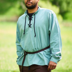 Op de afbeelding: Een man draagt een lichtblauw, lang mouwig shirt met een zwarte veterkraag en een bruine leren riem. Hij staat in een grasveld.