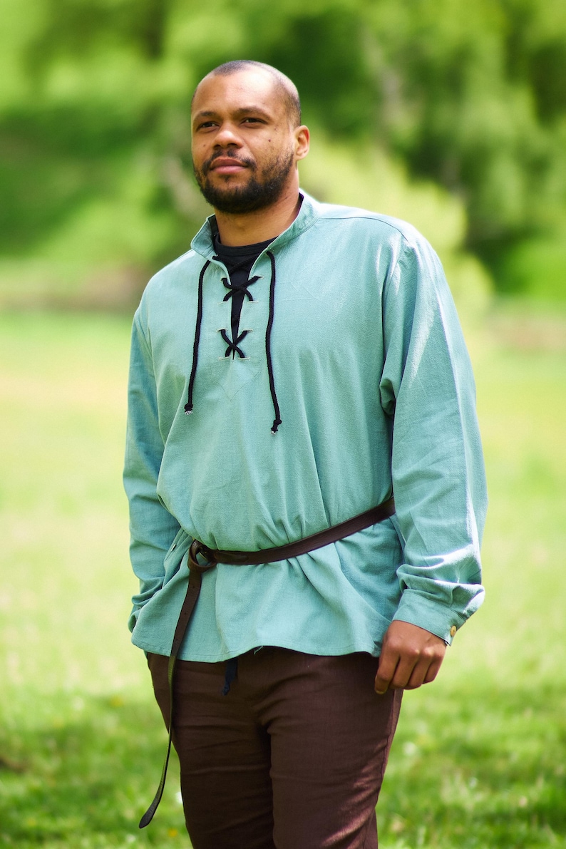 Op de afbeelding: Een man draagt een lichtblauw, lang mouwig shirt met een zwarte veterkraag en een bruine leren riem. Hij staat in een grasveld.