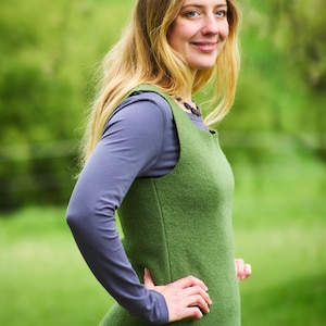 May include: A woman wearing a green sleeveless vest over a gray long-sleeved shirt. She is standing in a grassy field and smiling at the camera.