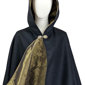 May include: A black hooded cape featuring a metallic olive green lining with a subtle floral pattern. The garment is secured at the neck with an ornate, filigree-style silver-toned metal clasp, creating a structured silhouette on a mannequin.