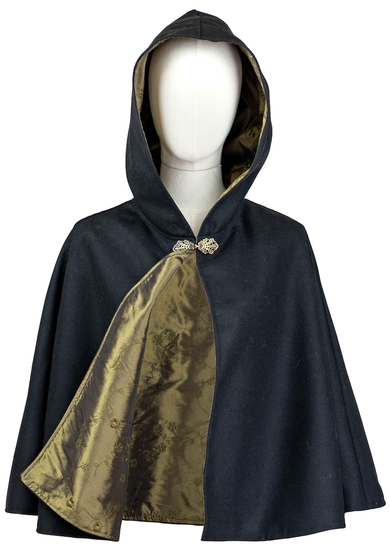 May include: A black hooded cape featuring a metallic olive green lining with a subtle floral pattern. The garment is secured at the neck with an ornate, filigree-style silver-toned metal clasp, creating a structured silhouette on a mannequin.