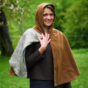 May include: A hooded cape featuring a solid brown exterior and a contrasting light green interior lining with a subtle floral pattern. The cape is secured at the neck with a single dark button and drapes over the shoulders.
