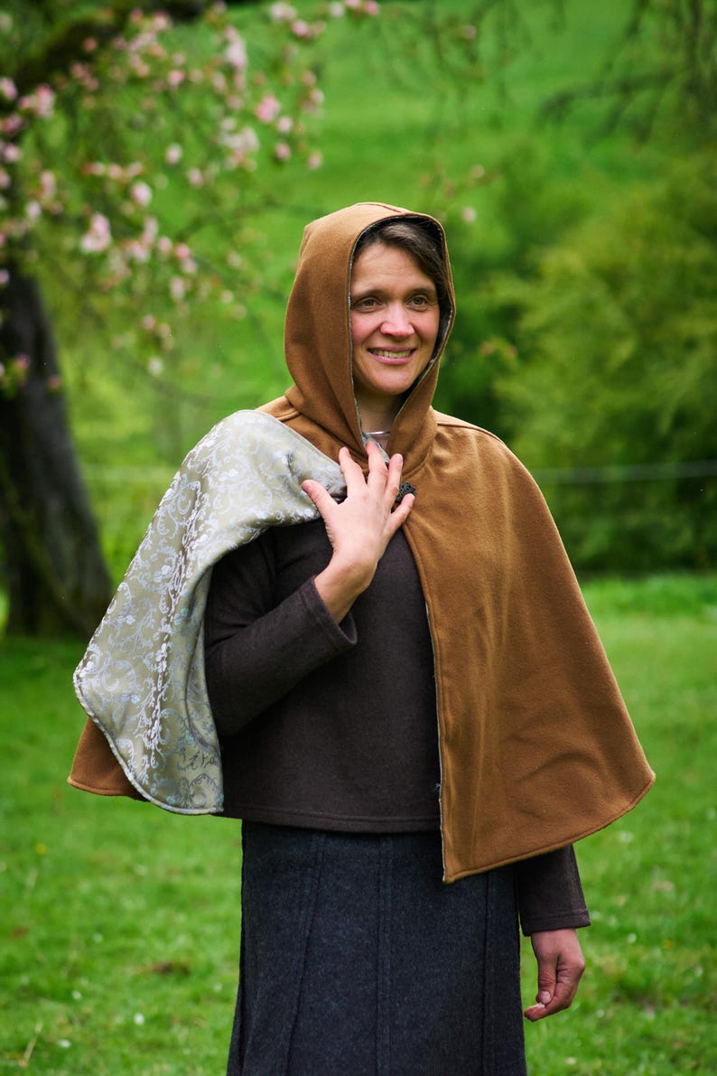 May include: A hooded cape featuring a solid brown exterior and a contrasting light green interior lining with a subtle floral pattern. The cape is secured at the neck with a single dark button and drapes over the shoulders.