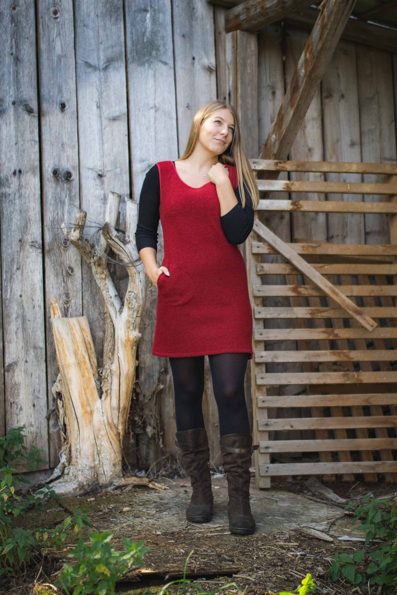 Puede incluir: Una mujer con un vestido rojo de punto con mangas negras y medias negras. Est&aacute; de pie frente a una pared de madera y una valla de madera. Lleva botas marrones.