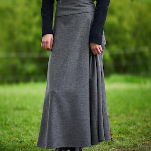 May include: A long, gray skirt with a high waist and a flowing silhouette. The skirt is paired with a black long-sleeved top. The outfit is worn outdoors on a grassy field. The skirt appears to be made of a textured fabric.