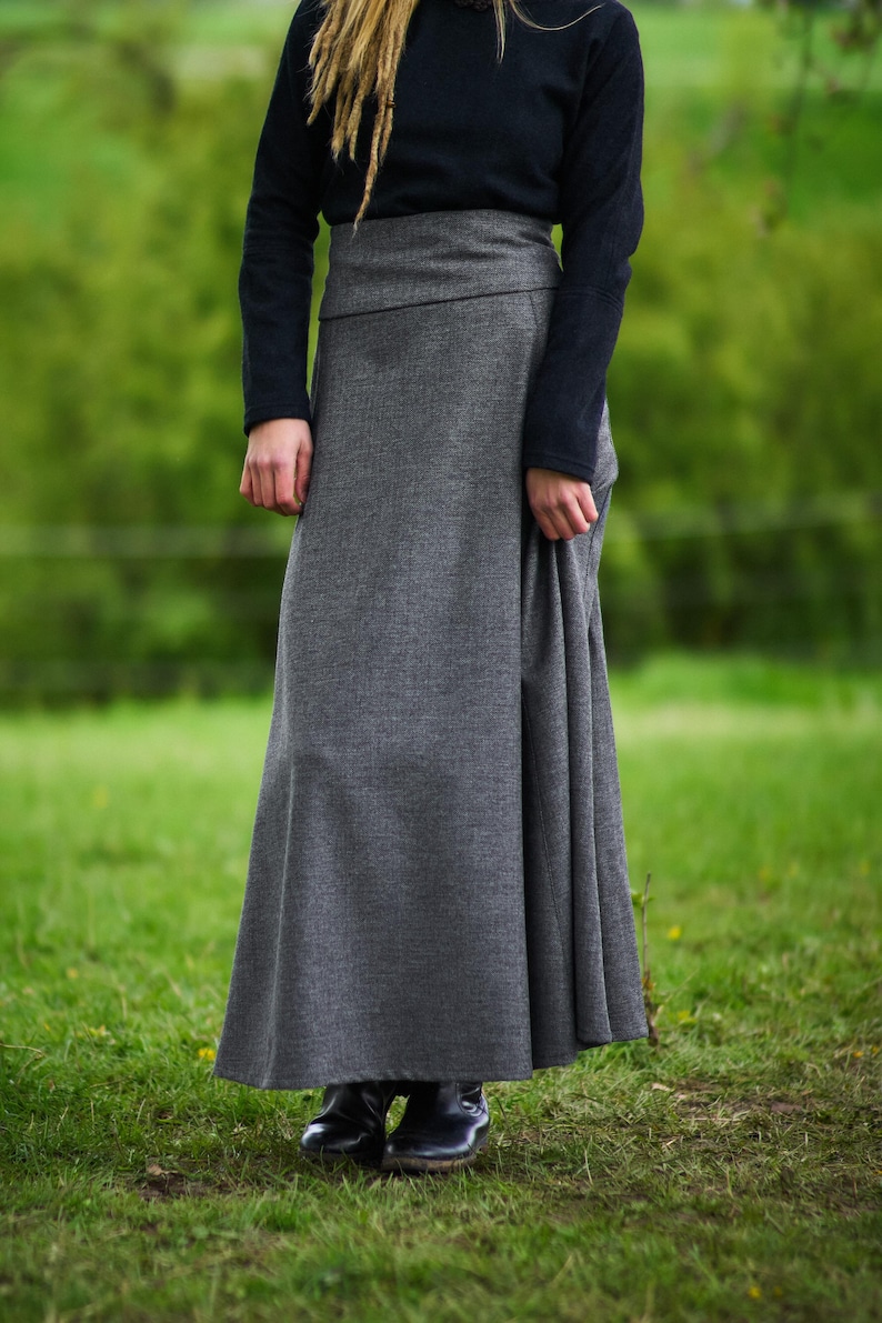 May include: A long, gray skirt with a high waist and a flowing silhouette. The skirt is paired with a black long-sleeved top. The outfit is worn outdoors on a grassy field. The skirt appears to be made of a textured fabric.
