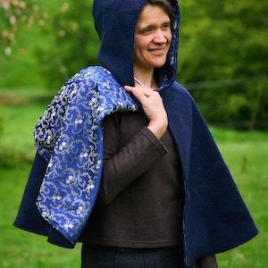 May include: A dark blue hooded cape featuring a vibrant royal blue lining with a detailed silver floral vine pattern. The outer layer is a solid dark blue fabric, and the garment is designed to drape over the shoulders with a hood.