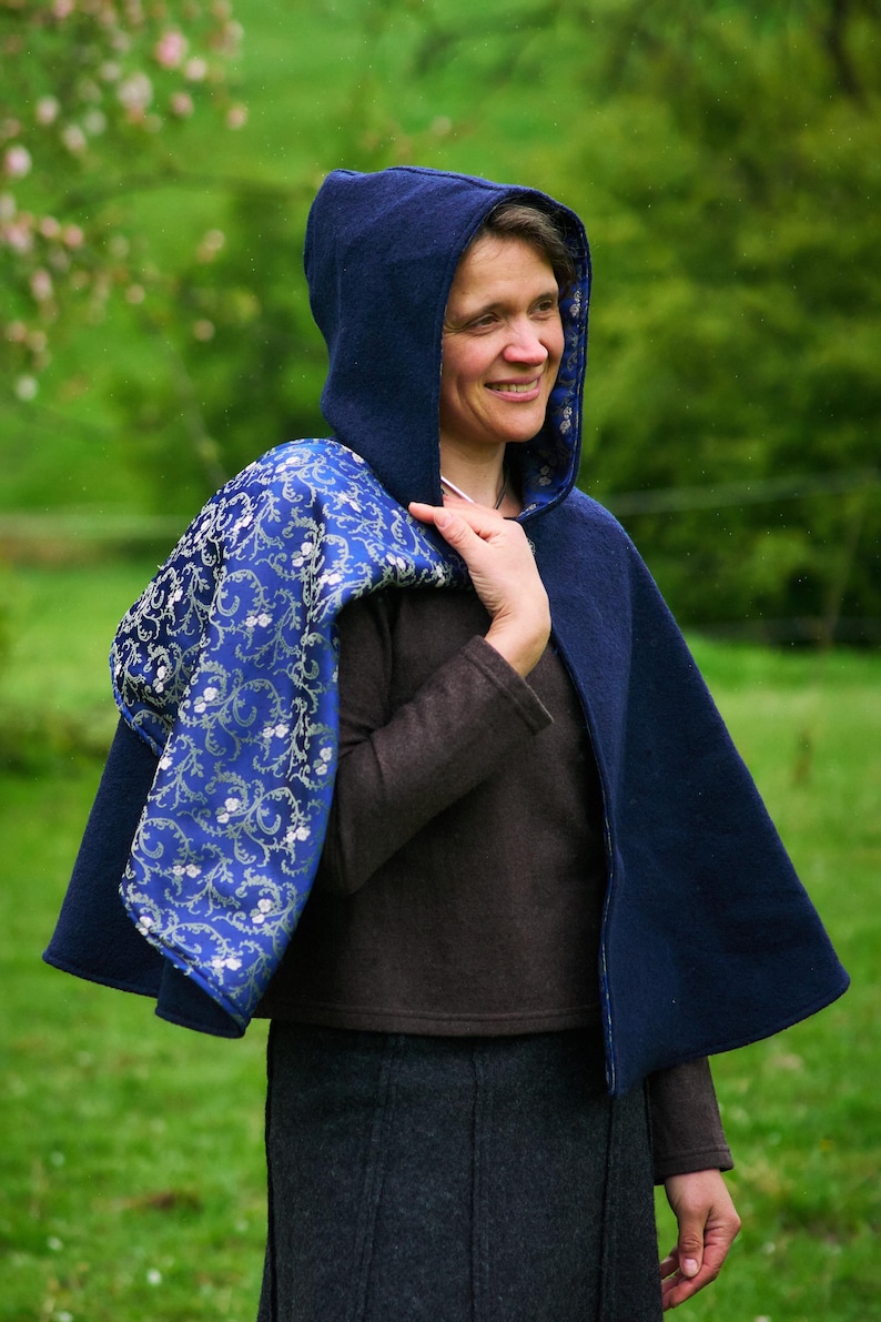 May include: A dark blue hooded cape featuring a vibrant royal blue lining with a detailed silver floral vine pattern. The outer layer is a solid dark blue fabric, and the garment is designed to drape over the shoulders with a hood.