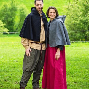 Pode incluir: Um casal posa num campo gramado, vestindo roupas de estilo medieval. A pessoa à esquerda usa uma capa preta com capuz, camisa bege, calças verde-oliva e botas marrons. A pessoa à direita usa uma capa cinza com capuz e uma saia longa vermelha.