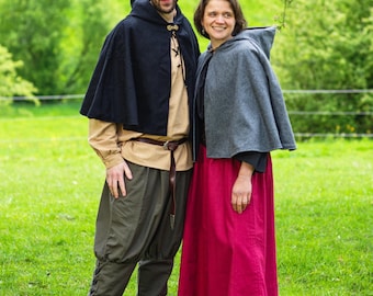 May include: A couple stands in a grassy field, wearing medieval-style clothing. The person on the left wears a black hooded cape, tan shirt, olive green pants, and brown boots. The person on the right wears a gray hooded cape and a long, red skirt.