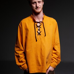 Op de afbeelding: Een man draagt een lang mouwig, oranje shirt met een zwarte veterkraag. Hij staat met zijn handen in zijn zakken en draagt een bruine broek.