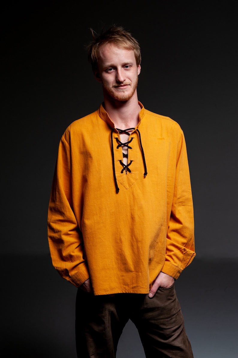 Op de afbeelding: Een man draagt een lang mouwig, oranje shirt met een zwarte veterkraag. Hij staat met zijn handen in zijn zakken en draagt een bruine broek.