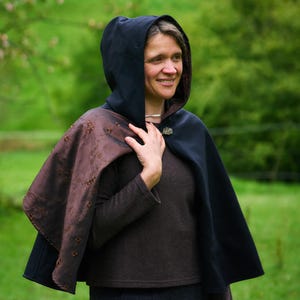 May include: A hooded cape featuring a solid black exterior and a contrasting dark brown interior lining with an embroidered floral pattern. The cape is secured at the neck with a decorative metal clasp and worn over a brown long-sleeve top.