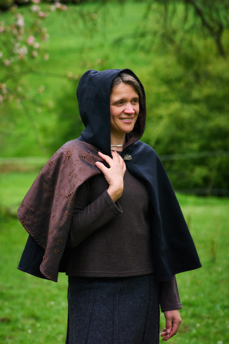 May include: A hooded cape featuring a solid black exterior and a contrasting dark brown interior lining with an embroidered floral pattern. The cape is secured at the neck with a decorative metal clasp and worn over a brown long-sleeve top.