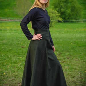May include: A woman wearing a dark gray long-sleeved top and a long, dark green skirt. The skirt has a high waist and a flowing A-line shape. The woman is standing in a grassy field with trees in the background.