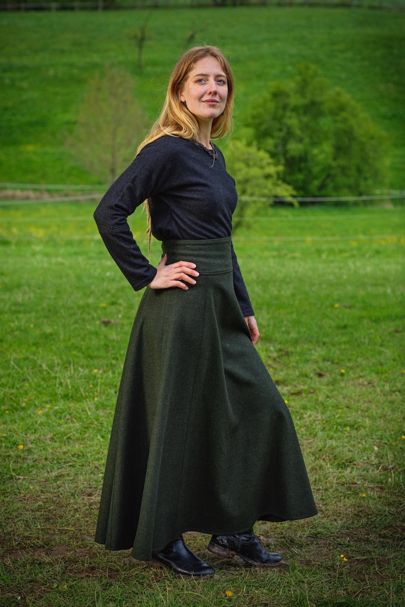 May include: A woman wearing a dark gray long-sleeved top and a long, dark green skirt. The skirt has a high waist and a flowing A-line shape. The woman is standing in a grassy field with trees in the background.