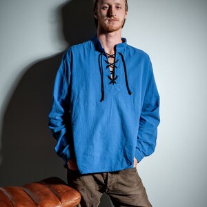 Op de afbeelding: Een man draagt een blauw, lang mouwig shirt met een veterkraag en een bruine broek. Hij staat naast een bruine leren kruk.