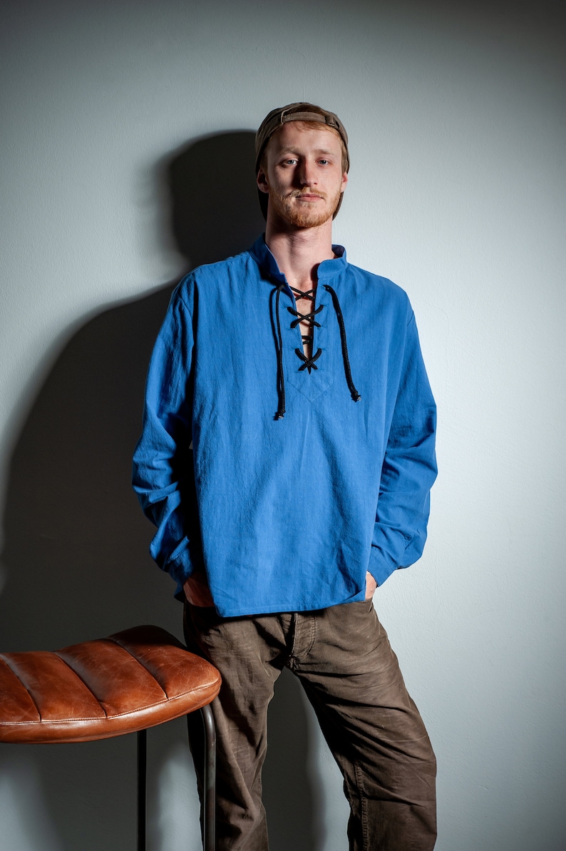 Op de afbeelding: Een man draagt een blauw, lang mouwig shirt met een veterkraag en een bruine broek. Hij staat naast een bruine leren kruk.