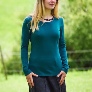 Könnte beinhalten: Eine Frau trägt ein türkisfarbenes Langarmshirt und einen dunkelgrauen Rock. Sie steht auf einer grünen Wiese.