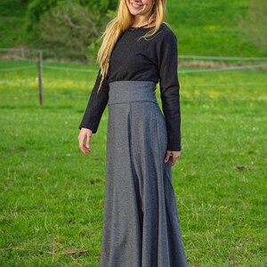 May include: A long-sleeved, dark gray top paired with a high-waisted, flowing gray skirt. The outfit is completed with black boots. The person is smiling in a grassy field, suggesting outdoor wear.