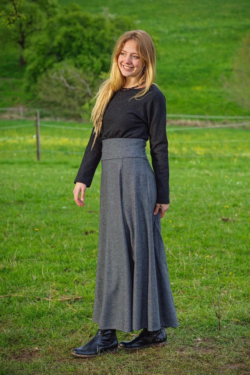 May include: A long-sleeved, dark gray top paired with a high-waisted, flowing gray skirt. The outfit is completed with black boots. The person is smiling in a grassy field, suggesting outdoor wear.