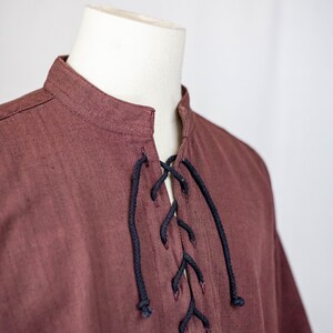 May include: A maroon shirt with a black lace-up neckline. The shirt has a mandarin collar and long sleeves.