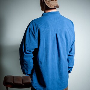 Op de afbeelding: Een persoon draagt een blauw lang mouw shirt met een bruine baseballpet met daarop "Sport". De persoon staat voor een lichtgrijze muur.