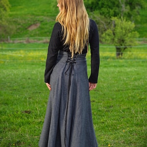 May include: A long, gray skirt with a high waist and a laced back detail. The skirt is paired with a black long-sleeved top. The person is standing in a grassy field with trees in the background.