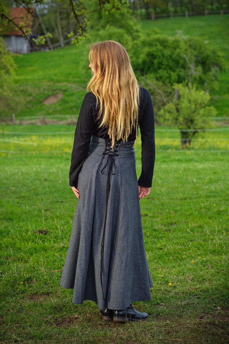 May include: A long, gray skirt with a high waist and a laced back detail. The skirt is paired with a black long-sleeved top. The person is standing in a grassy field with trees in the background.