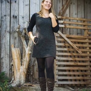 Puede incluir: Una mujer con un vestido gris, medias negras y botas marrones, de pie frente a una pared y una valla de madera.