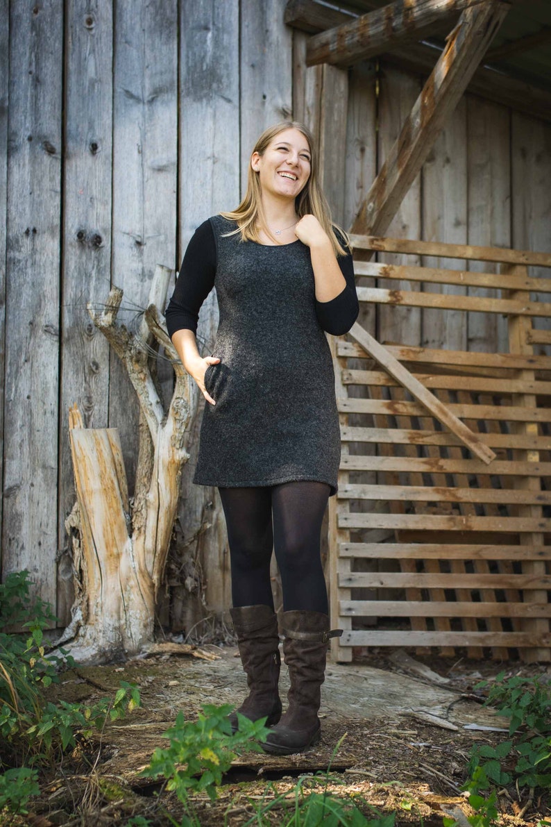 Puede incluir: Una mujer con un vestido gris, medias negras y botas marrones, de pie frente a una pared y una valla de madera.