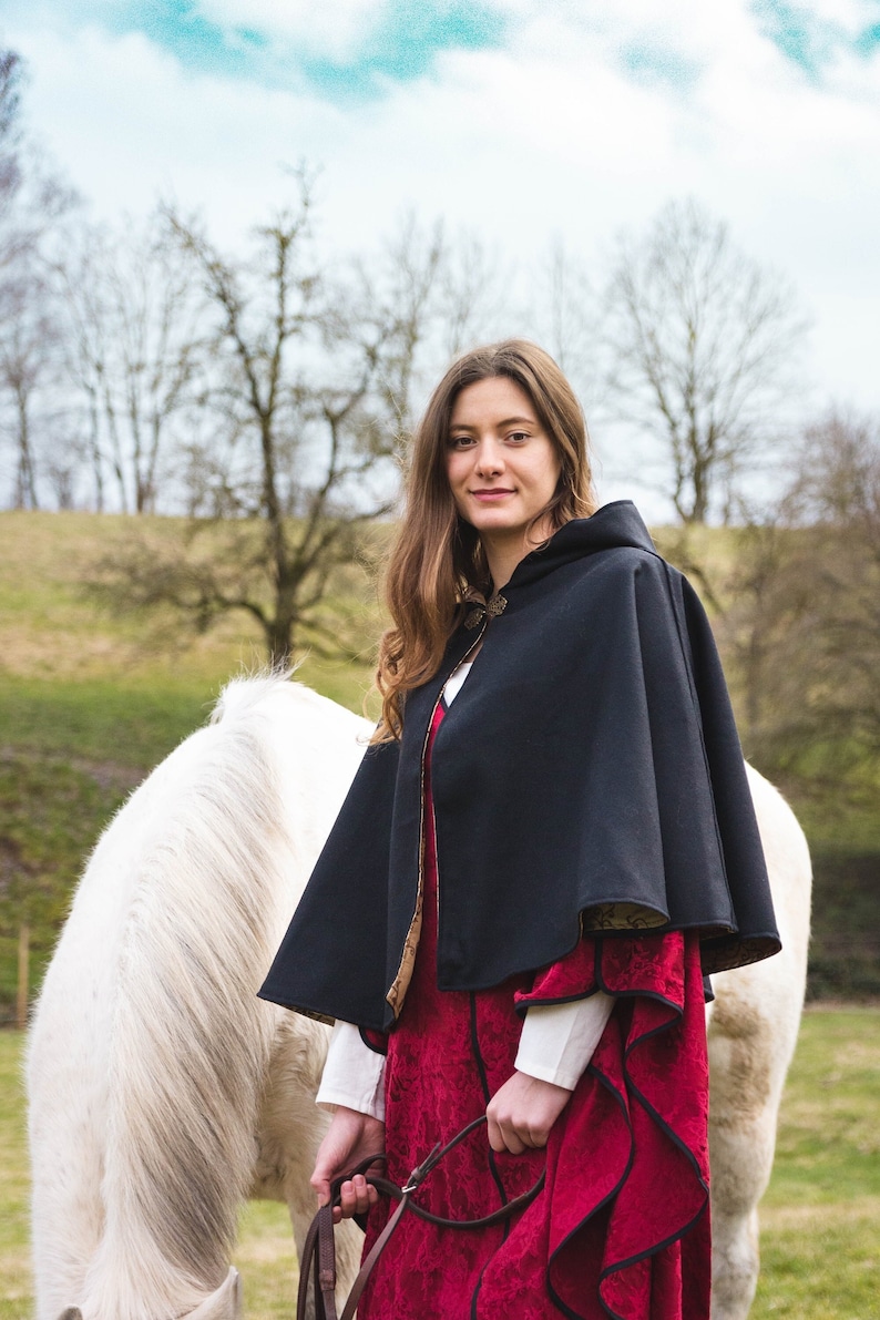 May include: A black hooded cloak with a patterned lining worn over a red dress with black trim and white sleeves. The cloak features a decorative metal clasp at the neckline. The garment is layered, with the red dress showing a textured floral pattern.