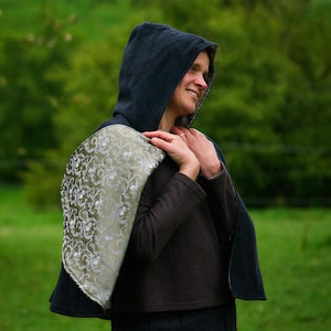 May include: A hooded cape featuring a dark charcoal exterior and a contrasting olive green lining with a silver floral pattern. The garment is worn over a dark brown long-sleeved top and a dark charcoal skirt, covering the shoulders and back.