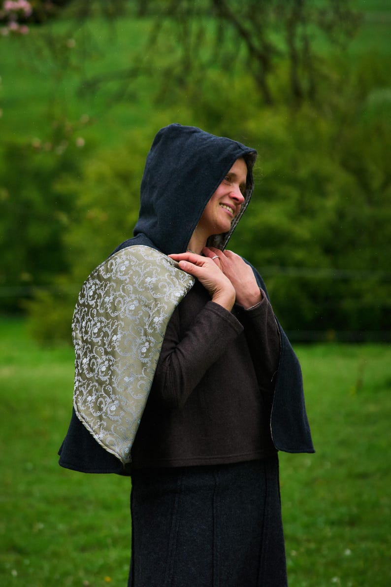 May include: A hooded cape featuring a dark charcoal exterior and a contrasting olive green lining with a silver floral pattern. The garment is worn over a dark brown long-sleeved top and a dark charcoal skirt, covering the shoulders and back.